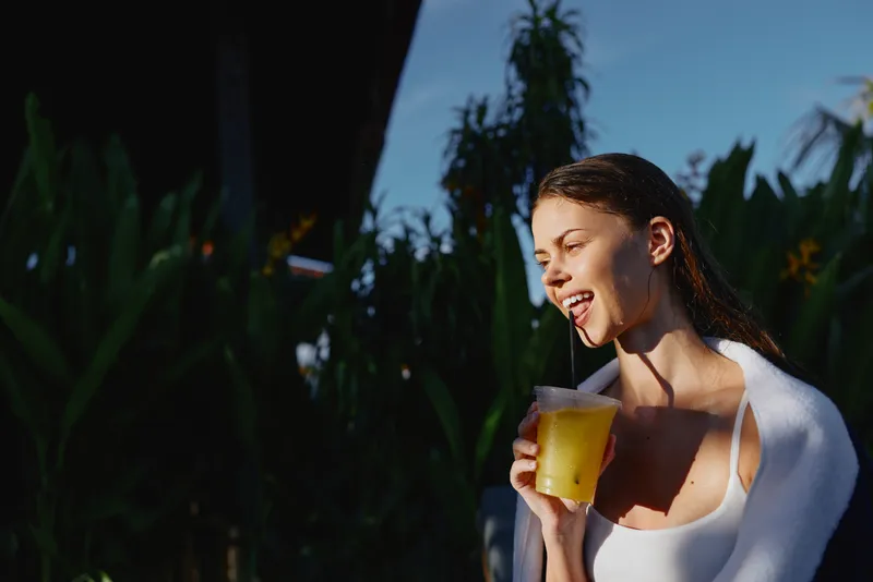 Young woman with wet hair enjoying a cold drink outdoors in natural sunlight. Relaxed lifestyle concept with tropical plants in the background and casual summer vibes.