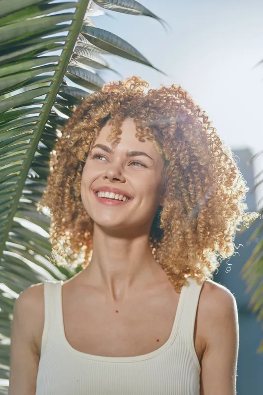 smiling woman with curly hair enjoys sunny day near tropical palm leaves, wearing white tank top, natural light highlights joyful outdoor mood