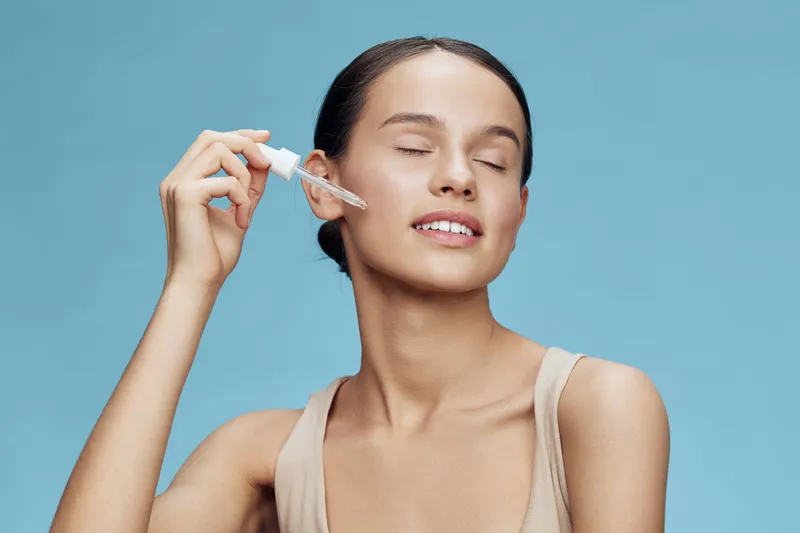 Young woman applying serum with dropper on glowing skin, wearing a beige top against a blue background, embodies beauty and skincare