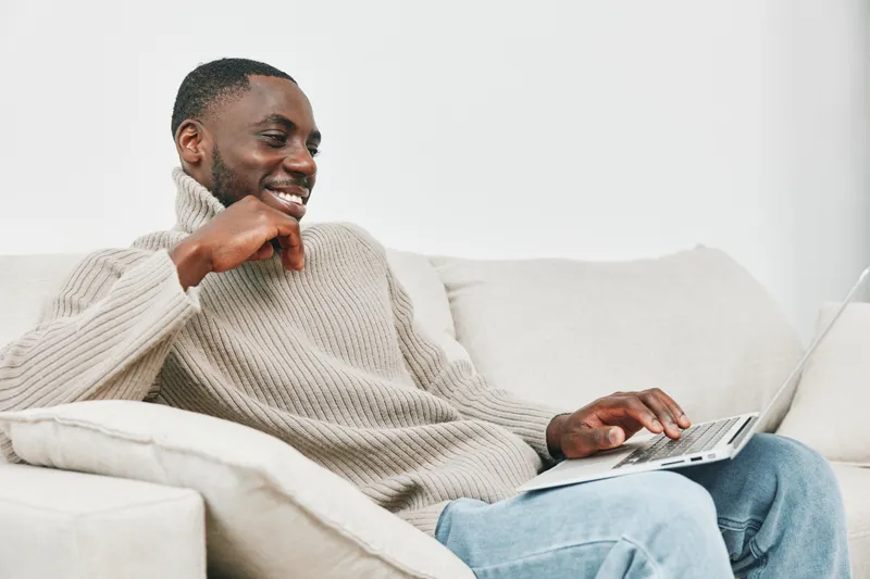 Smiling young man in a cozy beige sweater using a laptop at home for remote work, showcasing positivity and comfort in a modern living space