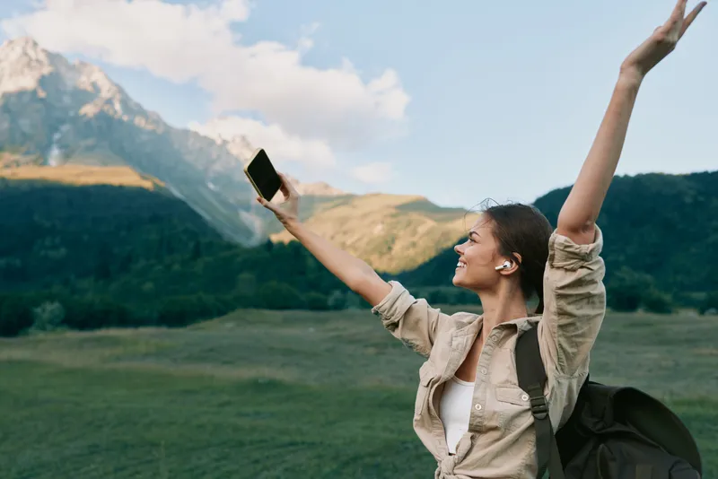 A happy woman in casual clothes raises her arms holding a smartphone in a mountainous landscape during daytime. Concept of freedom, lifestyle, and outdoor adventure.