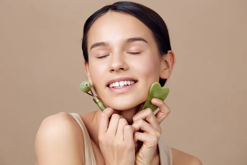 Young woman with flawless skin happily using jade roller and gua sha tool for facial massage in a neutral toned background, promoting natural beauty and skincare