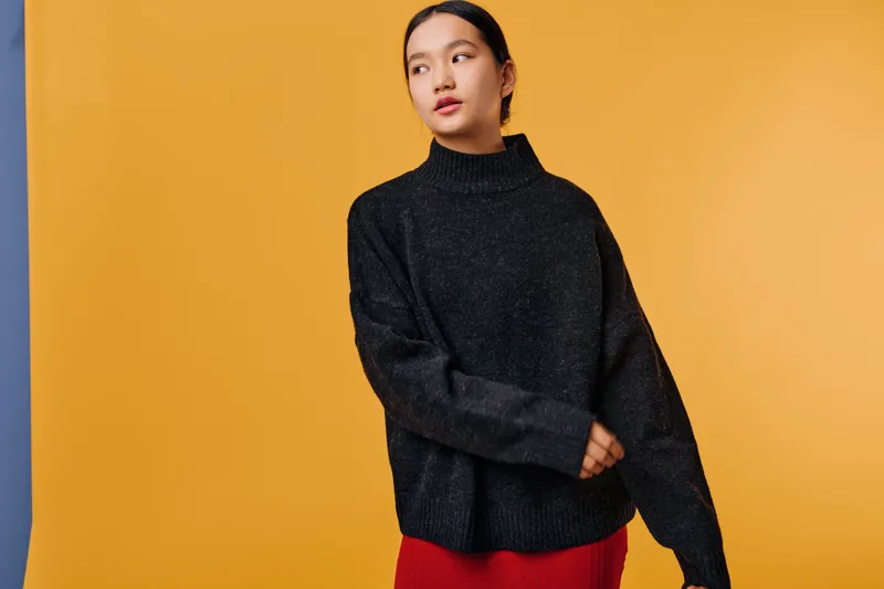 Woman wearing black oversized sweater and red skirt standing against orange background looking sideways with calm expression and casual stylish outfit for autumn fashion.