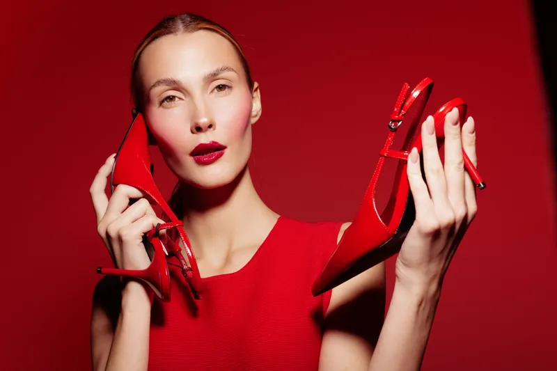 Fashion model in red dress holds red high heel shoes near face on red background. Concept of style, elegance, beauty, and modern fashion accessories for women.