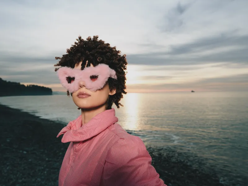 Woman wearing a pink mask at a sunset beach, posing for a portrait with coastal scenery, outdoor fashion mood, soft light, chic accessory styling by the seaside, calm water and horizon glow, stylish
