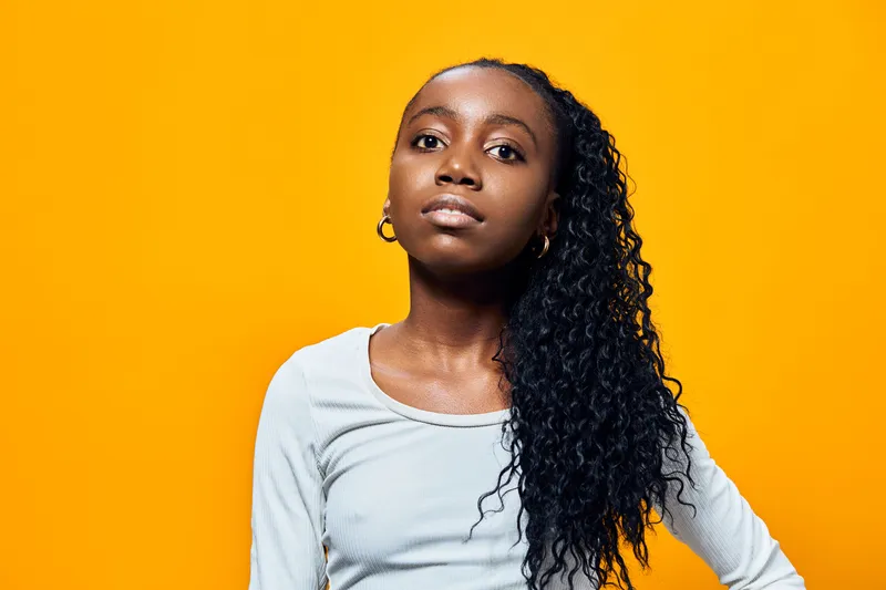 Confident young girl with curly hair posing against a vibrant yellow background, wearing a light gray long sleeve shirt, exuding strength and self assurance