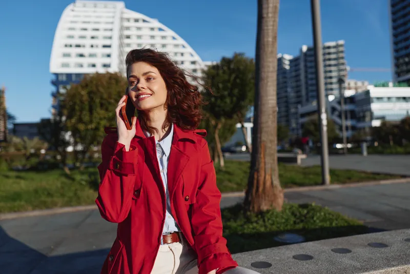 Young woman with brunette hair in a red trench coat talks on her smartphone while strolling in a sunny urban park with modern buildings in the background
