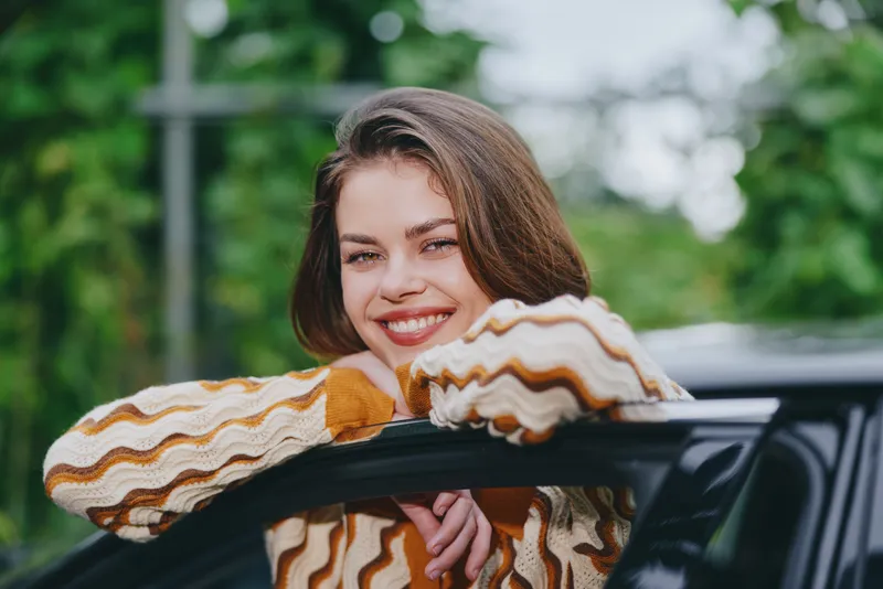 Cheerful woman leaning on a car window, smiling warmly in a casual striped sweater, outdoor setting with lush greenery and soft daylight.