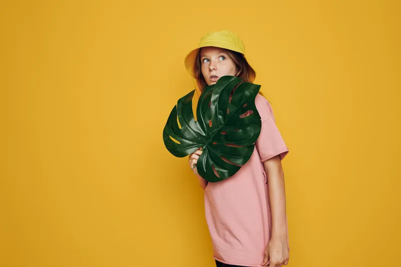 Young girl wearing a yellow hat and pink t-shirt holds a large green tropical leaf. She looks to the side with a curious expression. Bright yellow background emphasizes summer concept.