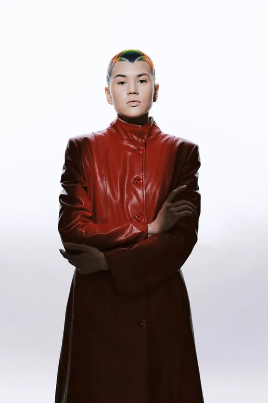Woman with short colorful hair wearing a red leather coat standing confidently with arms crossed against a bright white background, modern and edgy fashion style.