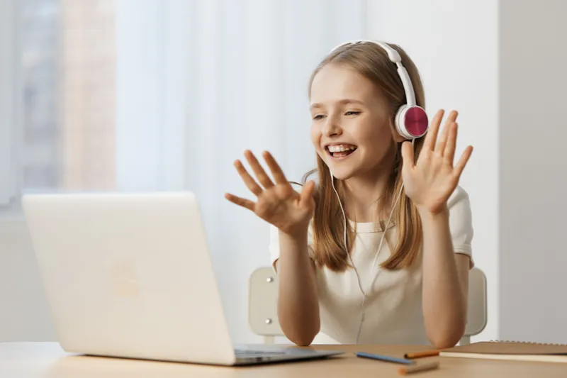 Cheerful girl with headphones smiling and waving while video chatting on a laptop, showcasing a bright and inviting workspace