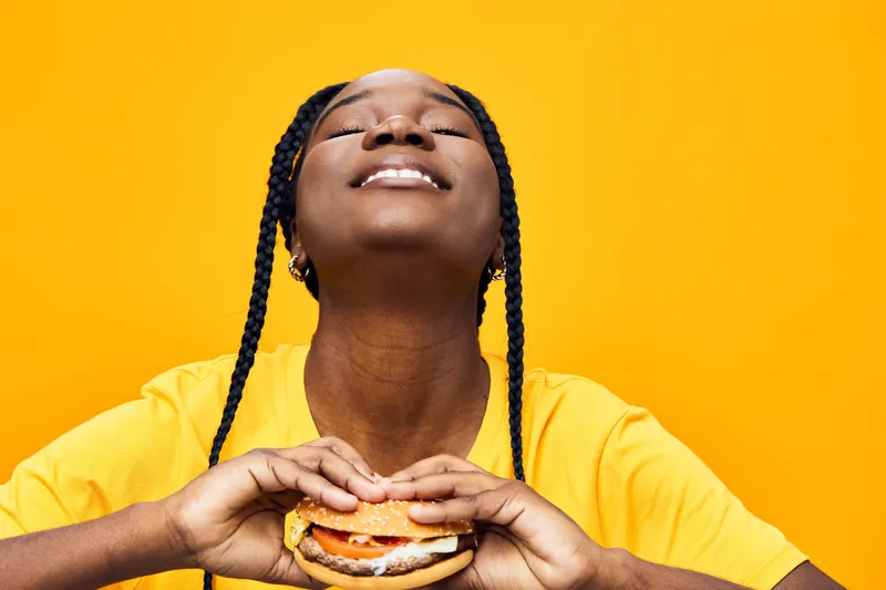 A joyful Black woman enjoying a delicious burger against a vibrant yellow background, reflecting happiness and indulgence in fast food