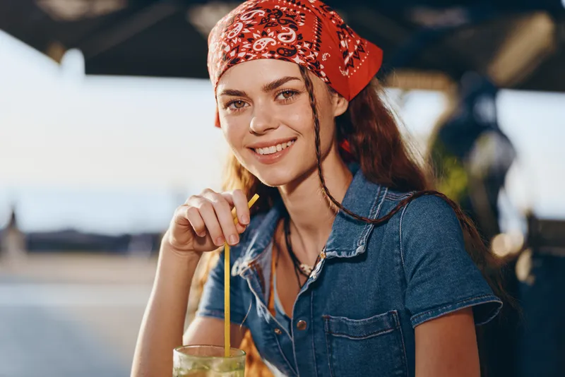 lifestyle woman in modern boho-western denim street style smiling with drink, film color, casual outdoor fashion, young female enjoying summer sunlight