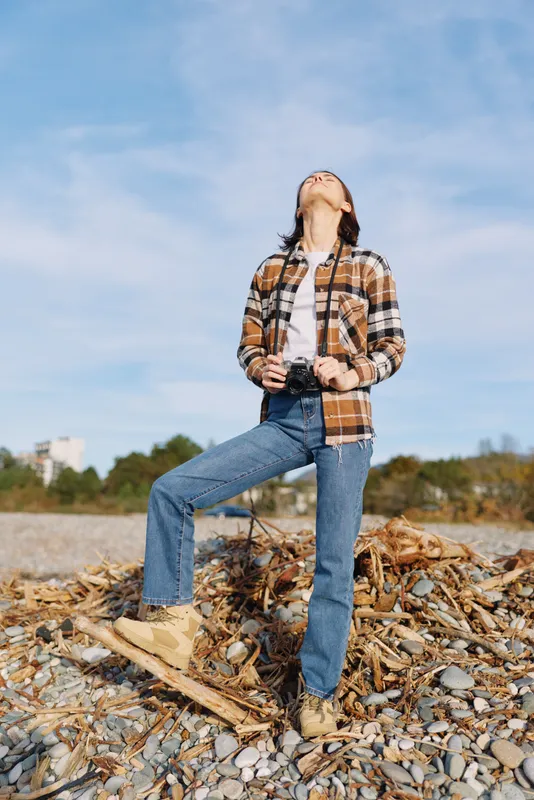Outdoor scene with a person in a plaid shirt and jeans on a pebbled beach with driftwood, looking up at a bright blue sky, casual fashion and outdoor exploration.