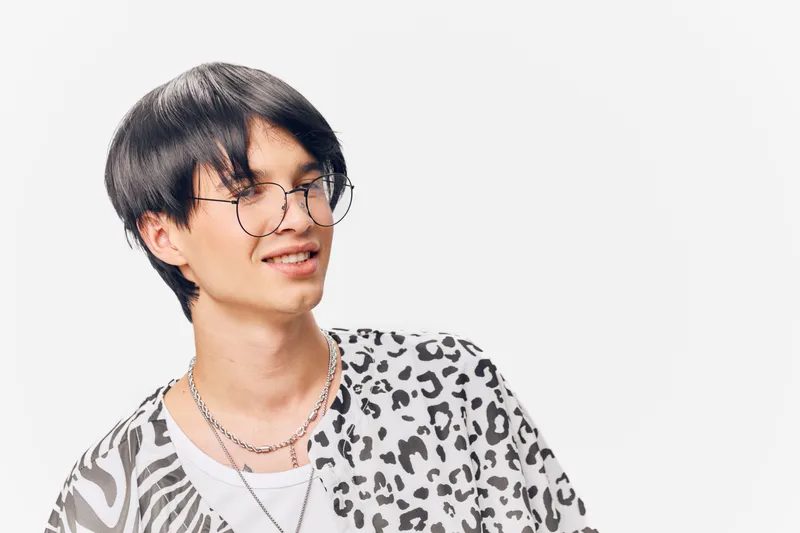 Young man wearing glasses and a stylish animal print shirt smiling confidently against a bright white background, conveying a modern casual vibe.