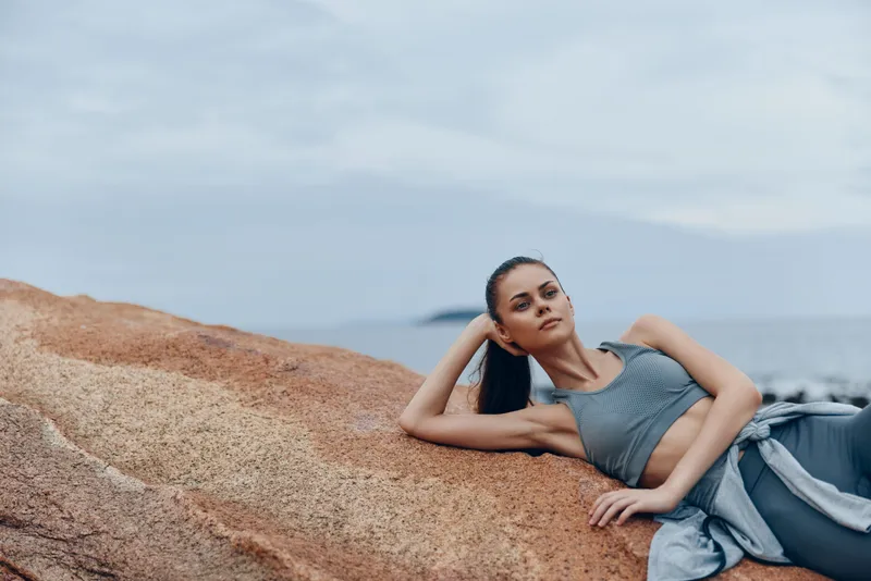 A fit young woman in activewear relaxing on a rock by the sea, showcasing a peaceful mood and a coastal background, perfect for fitness and wellness themes