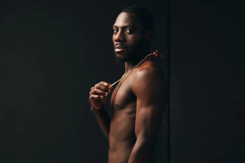 Creative conceptual portrait of a shirtless African man holding a flower with dark background, artistic visual style, strong expression and natural lighting in studio.