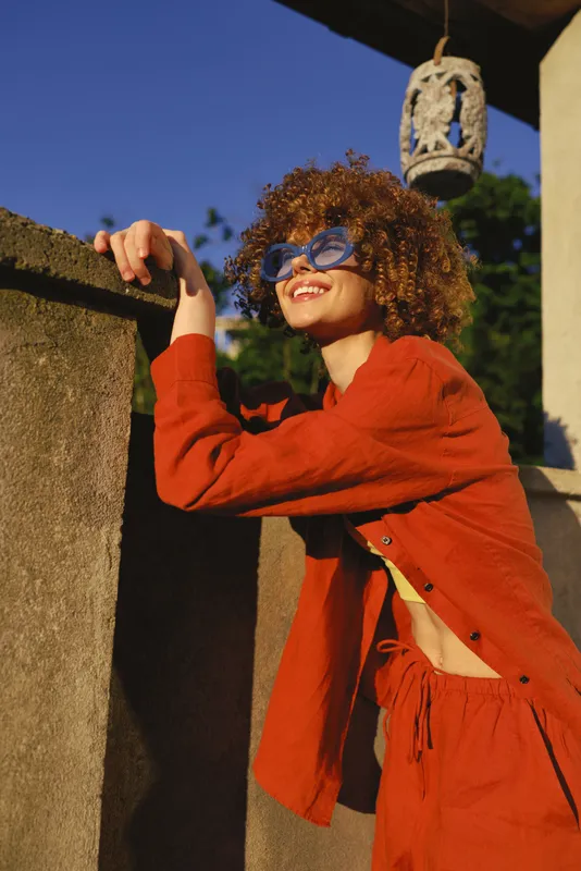 Cheerful woman outdoors, leaning on a wall, red cardigan, curly hair, blue glasses, sunlit moment, relaxed posture, warm tones, natural backdrop