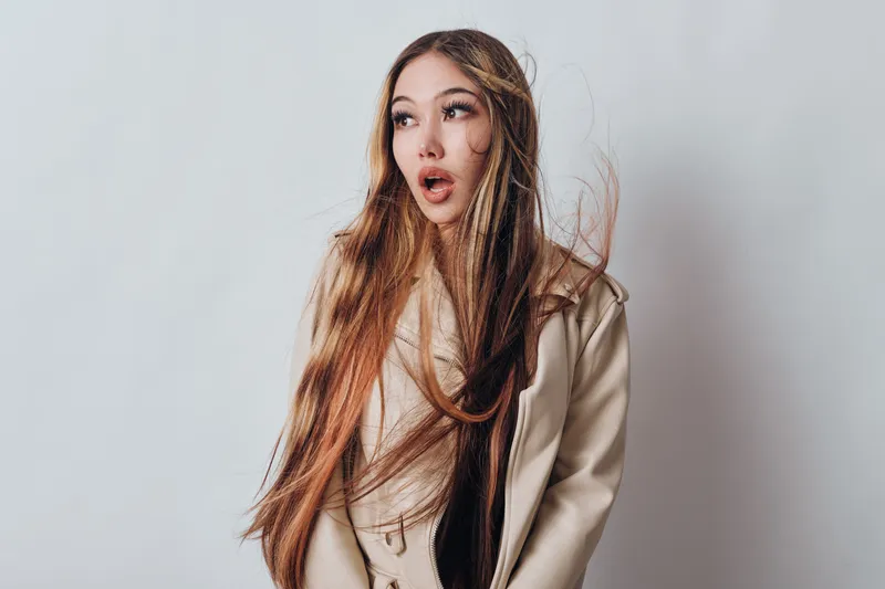 Young woman with long hair wearing beige jacket on light background looking surprised and expressive for fashion portrait and beauty concept in studio.