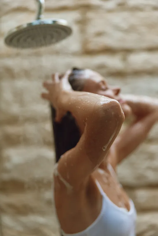 woman taking shower under rainfall showerhead with blurred stone wall background, wet skin, relaxed mood, natural light, warm color tones