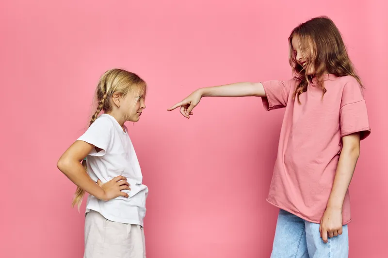 Two young girls facing each other on a pink background. One girl is pointing her finger while the other looks annoyed with hands on hips. Concept of conflict, argument.