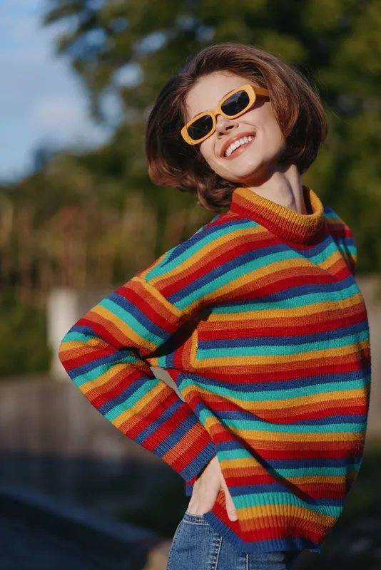 Woman smiling in a colorful rainbow sweater and yellow sunglasses outdoors, casual outfit with short hair, posing confidently on a sunny day for vibrant lifestyle and fashion portrait.