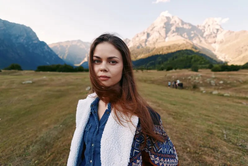 Woman portrait in front of mountains and meadow, nature outdoor travel countryside scene with long hair, casual jacket and serene expression, scenic landscape and distant peaks.