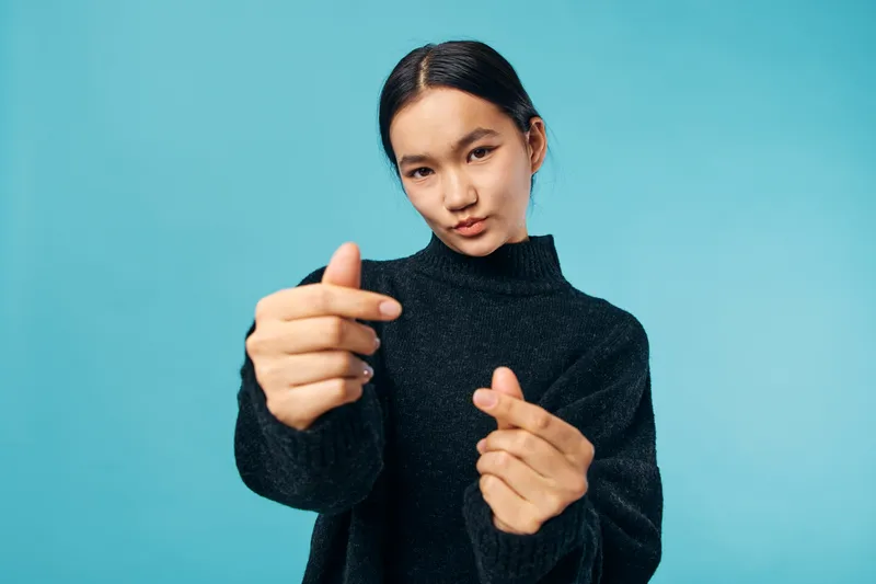 Young woman in black sweater posing with hand gestures for heart shape against light blue background in studio. Casual fashion, portrait and expression with clean colorful backdrop.