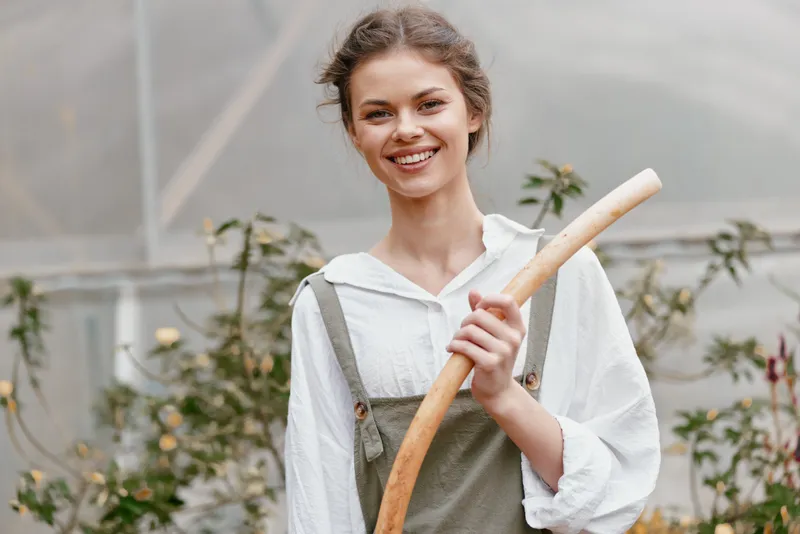 Young woman in overalls smiling in a greenhouse, holding a gardening tool, showcasing a connection to nature and healthy living, ideal for lifestyle concepts