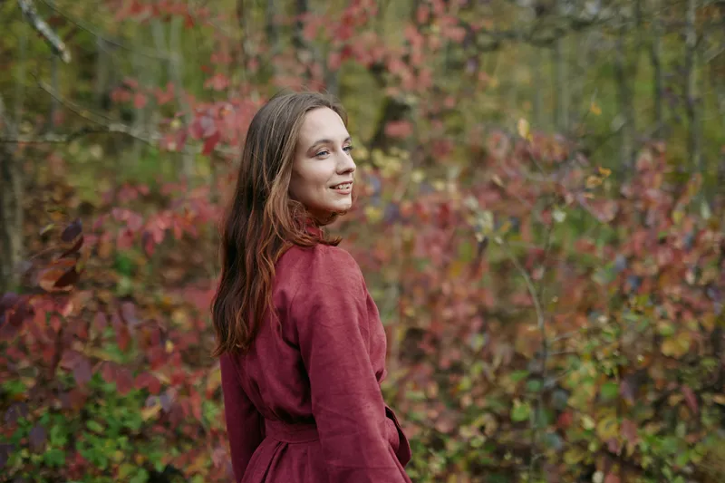 Candid portrait of a woman in a burgundy coat turning toward the camera in an autumn woodland, conveying natural authenticity and a genuine presence bathed in soft, warm light.