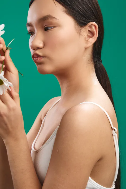 Asian woman in a light tank top softly smells a flower against a vivid green backdrop, creating a calm, intimate studio moment that highlights grace and natural charm.