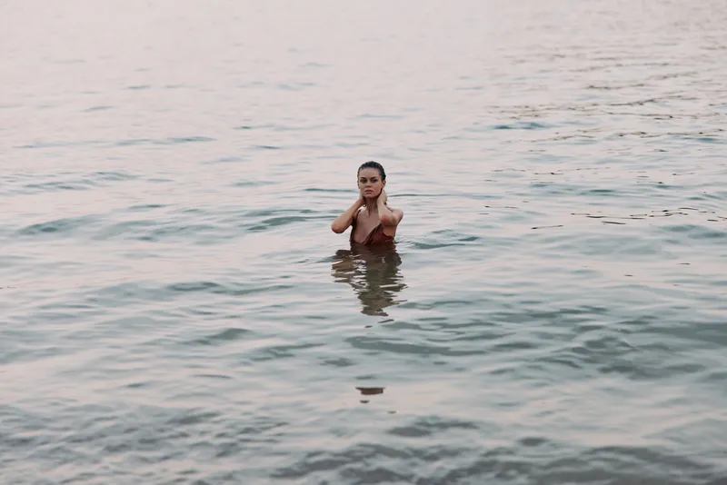 A solitary figure stands waist-deep in calm sea, hands near neck, gentle ripples surround, warm sunset light softly illuminates water and skin, creating a serene aquatic portrait.