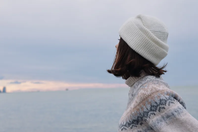 A person wearing a knit sweater and beanie stands by the sea, gazing toward the horizon. Winter mood, calm coastline, soft light, outdoor portrait by the water, quiet mood.