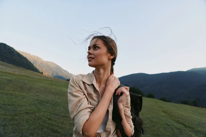 Cheerful woman outdoors with a backpack in a mountain meadow, sunlit breeze, casual jacket, relaxed posture, natural colors, showcasing a fresh outdoor lifestyle moment