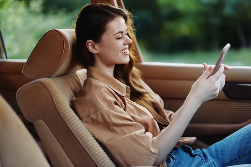 Young woman smiling while using smartphone in a car, wearing a casual brown shirt, green background, representing happiness and leisure.
