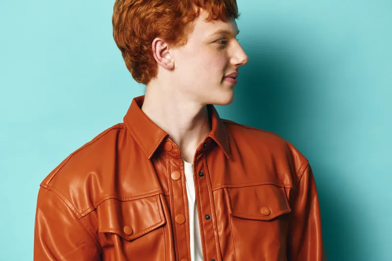 Young man with red hair wears brown leather jacket looking sideways on blue background. Casual fashion style, profile portrait and confident look closeup with studio lighting and clean backdrop.