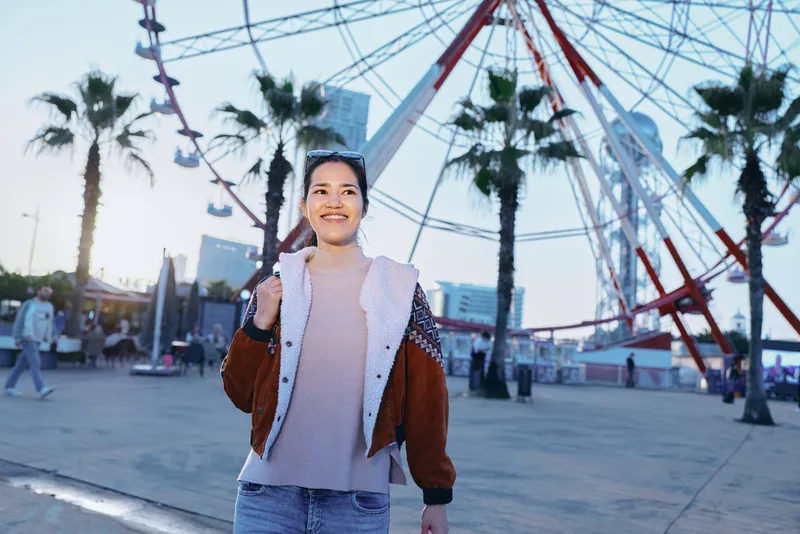 Woman enjoying outdoor amusement park with ferris wheel in the background, casual jacket and warm smile during sunset, capturing leisure, exploration and youthfulness