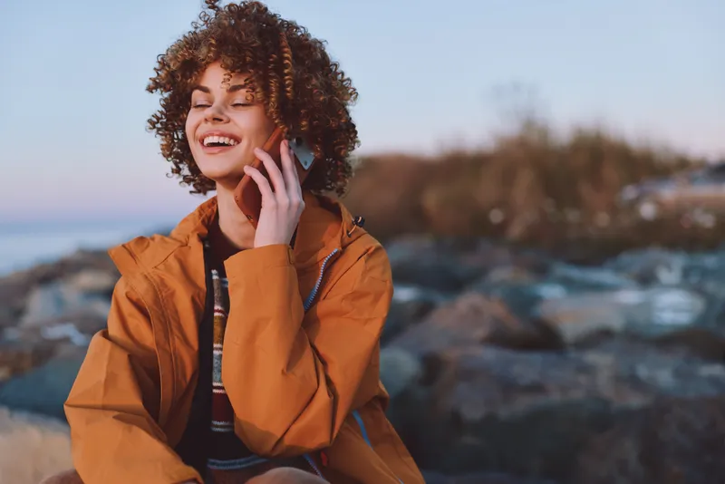 Woman curly hair smiling phone call outdoor lifestyle coast scene, sitting outdoors on rocky ground. Warm daylight and blurred sea create relaxed summer mood, candid portrait today.