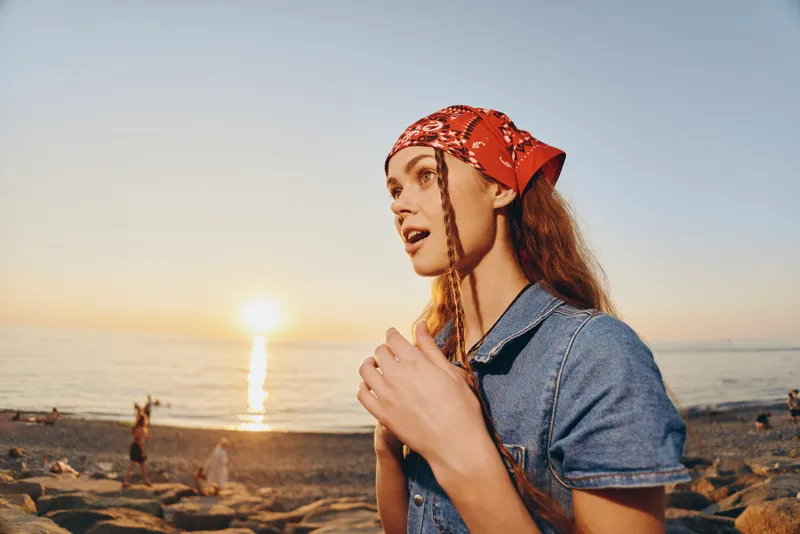 Lifestyle woman in modern boho-western denim street style gazes at sunset by the beach with warm film color tones creating a relaxed and natural mood.