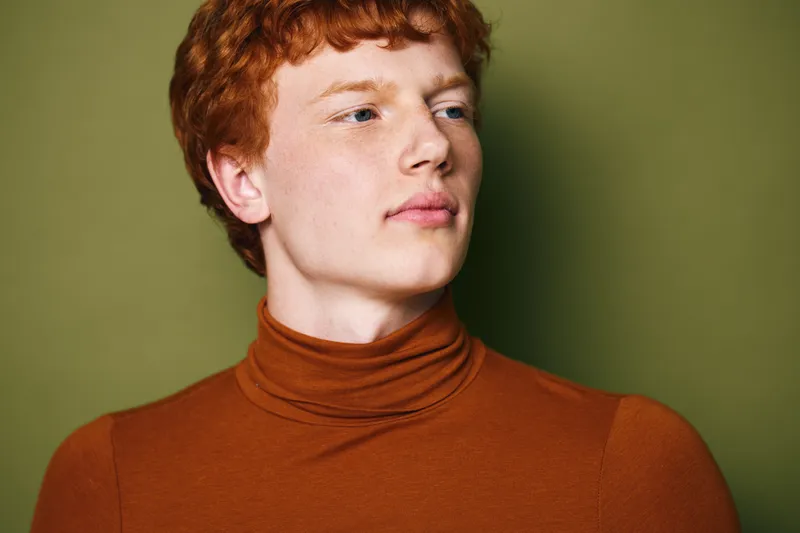 Young man with red hair wearing brown turtleneck posing on green background, portrait of thoughtful male model looking away with soft natural light and calm expression.