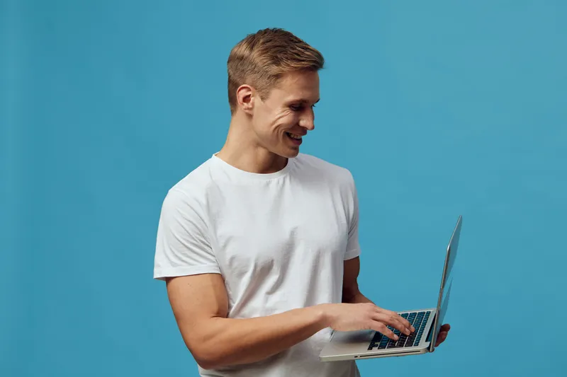 Smiling man holding a laptop computer, wearing a plain white t-shirt, standing against a bright turquoise background in a studio shoot. Casual technology lifestyle concept for communication and remote