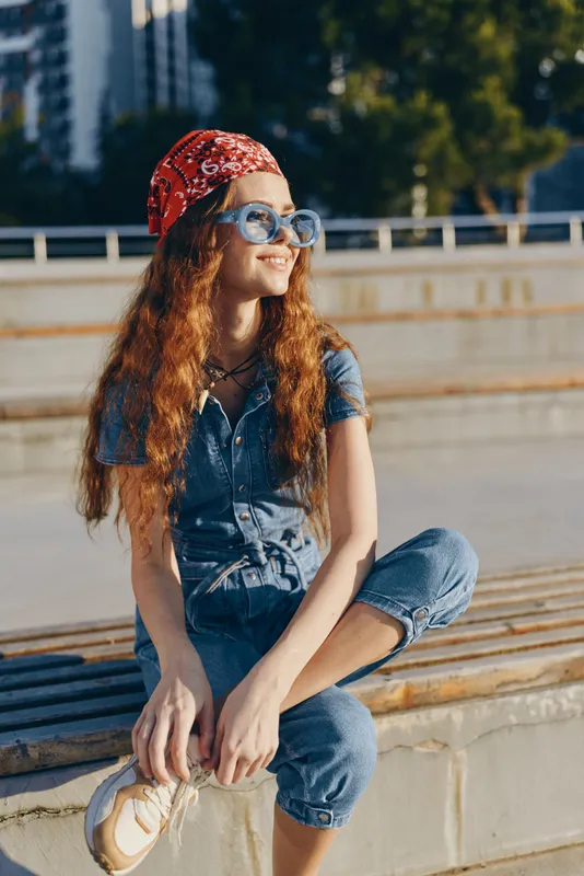 Lifestyle woman in modern boho-western denim street style sits relaxed outdoors wearing sunglasses and a red bandana, captured in warm film color tones.