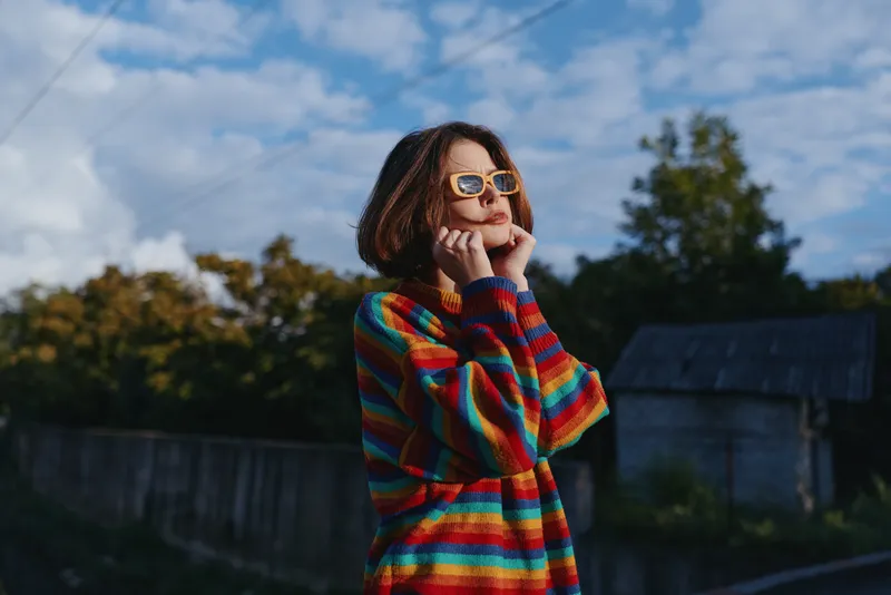 Woman in rainbow sweater and sunglasses smiling outdoors in a bright portrait, casual fashion and joyful expression with blue sky, trees and rustic fence in background.