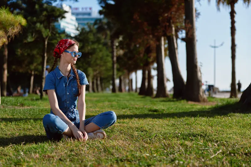 lifestyle woman in modern boho-western denim street style sitting casually on grass with film color tones, relaxed expression, sunny day outdoors