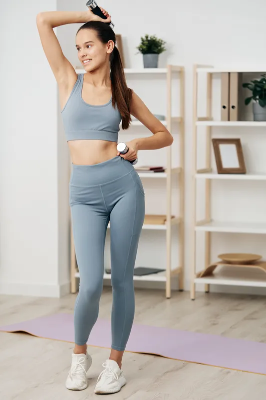 Young woman fitness athlete exercising indoors in matching grey sports bra and high waisted leggings, holding a dumbbell and water bottle on a yoga mat in bright home gym.