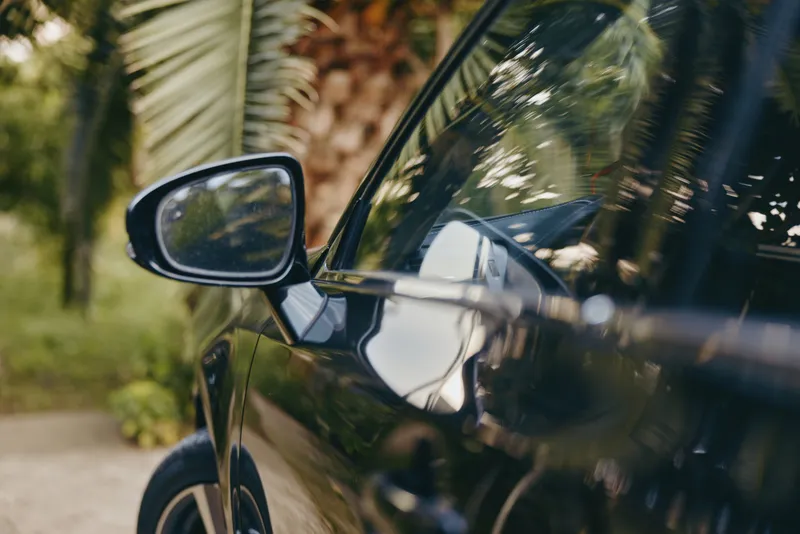 Black car side mirror and door handle with palm reflection in tropical outdoor setting, glossy paint on modern sedan conveys travel lifestyle and polished automotive detail.