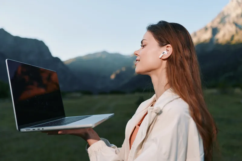 A cheerful professional woman uses a laptop outdoors in a scenic mountain setting. She wears light attire and looks focused, creating a calm, productive atmosphere and a sense of modern remote work.