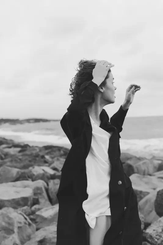 Woman standing by the sea on a windy day, hair and coat tossed by the breeze, looking toward the horizon. Black and white mood highlights coastal freedom, reflection, and style, suitable for fashion,