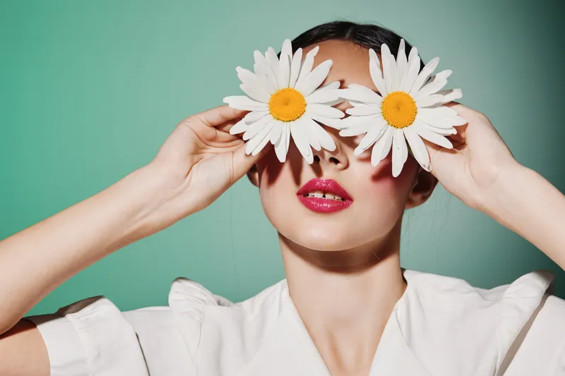 Young woman with glossy lips holding two large white flowers over eyes. Creative beauty and fashion concept on a pastel green background, styled for artistic editorial shoots.