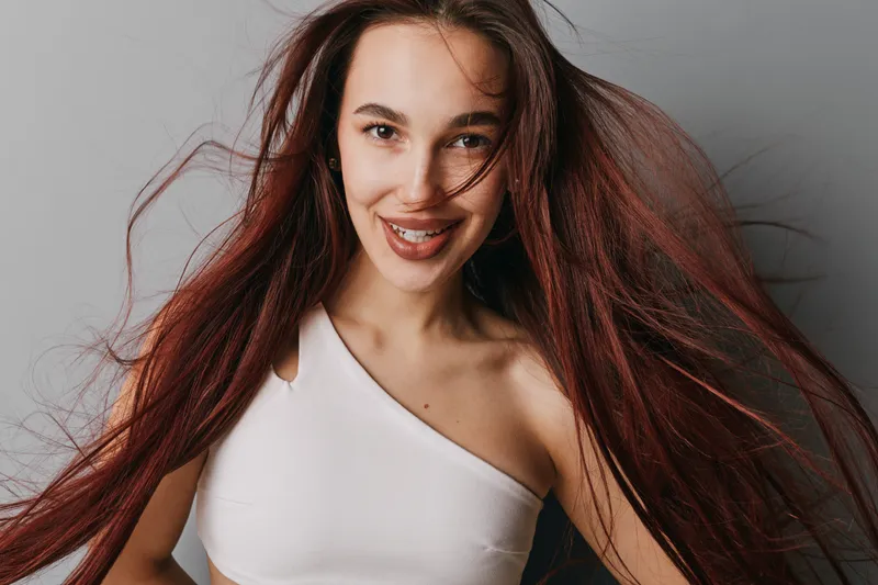 Young woman with long red hair smiling and posing in white top against gray background, natural beauty, confident expression and casual style in studio portrait photography.